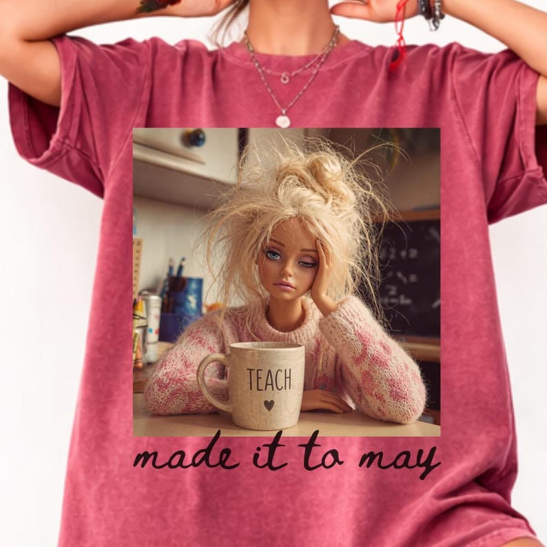 K&ouml;nnte beinhalten: Burgunderfarbenes T-Shirt mit einem aufgedruckten Bild einer Puppe mit zerzaustem blondem Haar, die ihren Kopf auf ihre Hand st&uuml;tzt. Sie tr&auml;gt einen rosa Pullover und h&auml;lt eine Tasse mit der Aufschrift "TEACH". Der Text "made it to may" ist darunter gedruckt.