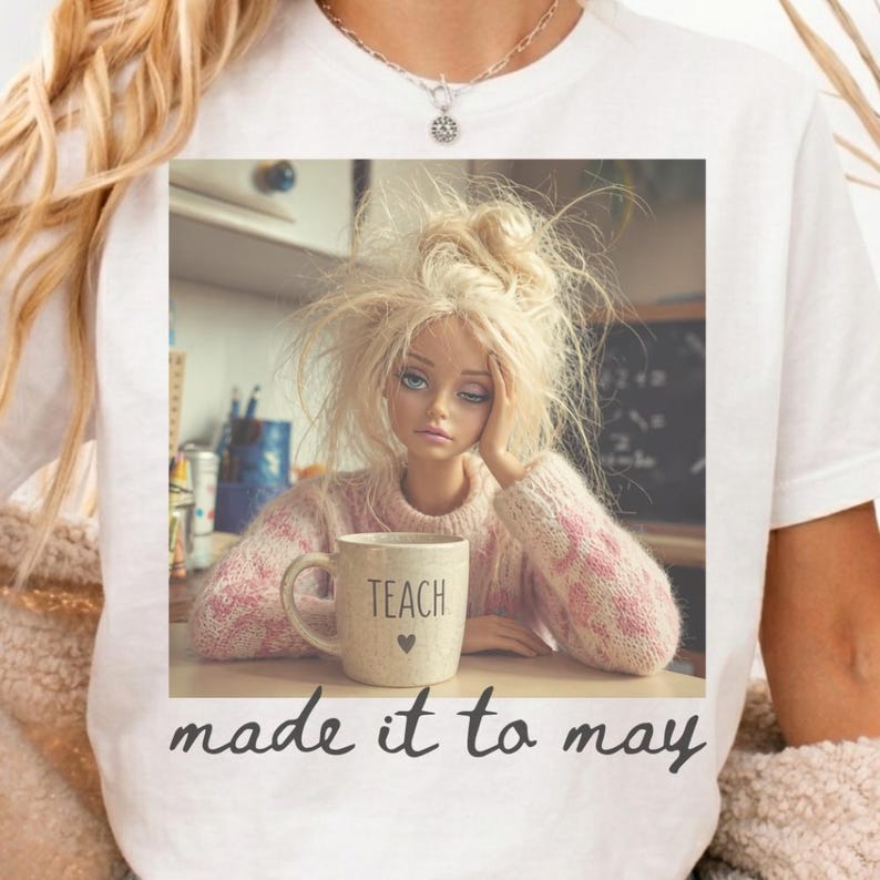 K&ouml;nnte beinhalten: Wei&szlig;es T-Shirt mit einer Puppe mit zerzaustem blondem Haar, die einen rosa Pullover tr&auml;gt und ihren Kopf auf ihre Hand st&uuml;tzt. Eine Tasse mit dem Wort "TEACH" und einem Herz befindet sich vor ihr. Der Text "made it to may" steht unten.