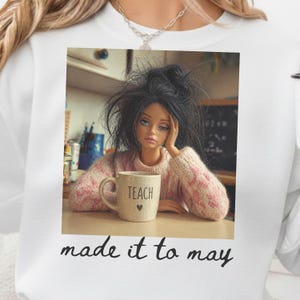 K&ouml;nnte beinhalten: Eine Puppe mit zerzaustem, schwarzem Haar und einem rosa-wei&szlig; gestreiften Pullover h&auml;lt eine Tasse mit der Aufschrift "TEACH" und einem Herz. Das Bild befindet sich in einem wei&szlig;en Sweatshirt mit den Worten "made it to may" am unteren Rand.