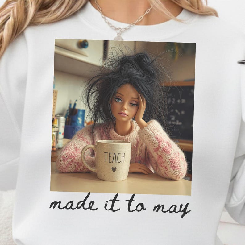 K&ouml;nnte beinhalten: Eine Puppe mit zerzaustem, schwarzem Haar und einem rosa-wei&szlig; gestreiften Pullover h&auml;lt eine Tasse mit der Aufschrift "TEACH" und einem Herz. Das Bild befindet sich in einem wei&szlig;en Sweatshirt mit den Worten "made it to may" am unteren Rand.