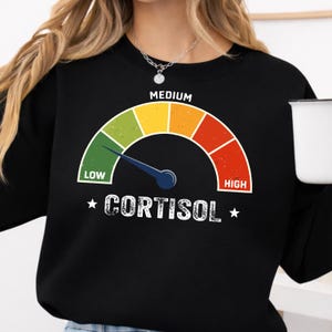 Könnte beinhalten: Schwarzes Sweatshirt mit einer Grafik eines Cortisolmessgeräts. Die Nadel des Messgeräts zeigt auf den grünen Bereich. Der Text "MEDIUM" steht oben, und "CORTISOL" ist darunter gedruckt.