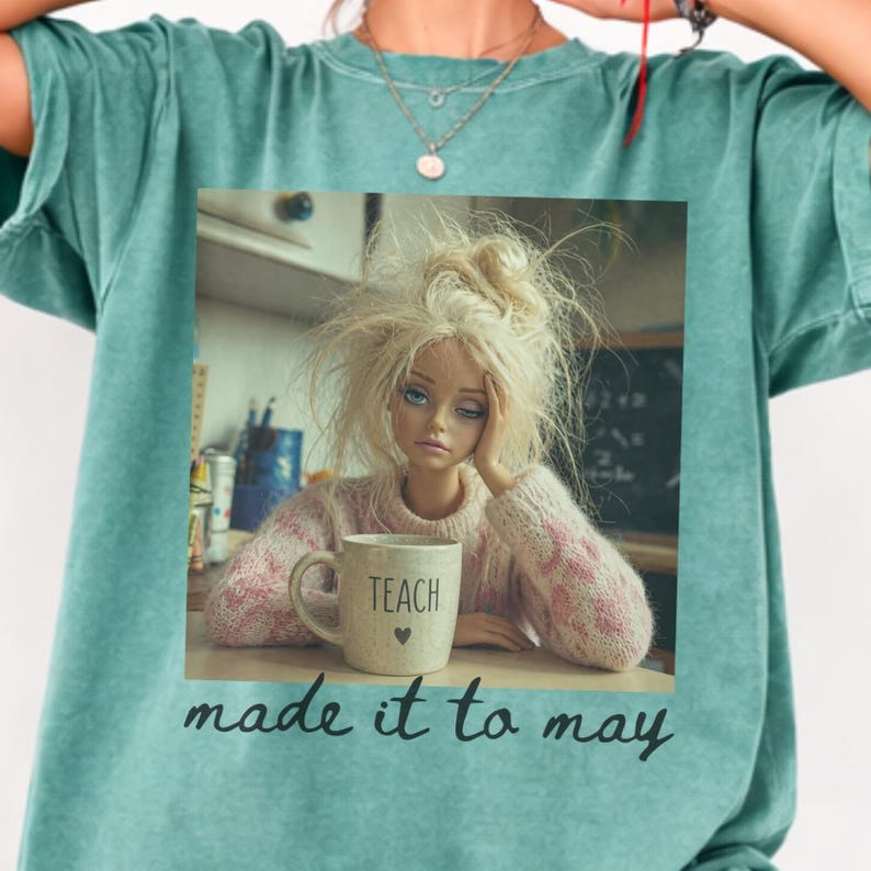 K&ouml;nnte beinhalten: T&uuml;rkisfarbenes T-Shirt mit einem quadratischen Bild einer Puppe mit zerzaustem blondem Haar, die einen rosa Pullover tr&auml;gt und eine Tasse mit der Aufschrift "TEACH" h&auml;lt. Der Text "made it to may" ist darunter gedruckt.