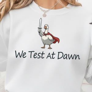 Może przedstawiać: Biała bluza z kapturem z kreskówkową gęsią w czerwonej pelerynie i trzymającą miecz. Tekst "We Test At Dawn" jest wydrukowany na czarno pod ilustracją gęsi. Bluza to swobodny, wygodny styl.