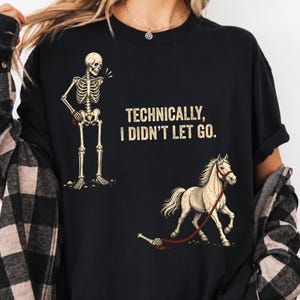 Puede incluir: Una camiseta negra con un gráfico de un esqueleto de pie a la izquierda y un caballo blanco a la derecha. El esqueleto sostiene una cuerda roja atada al caballo, con una mano esquelética separada que aún sujeta la cuerda. El texto dice: TECHNICALLY, I DIDN'T LET GO.