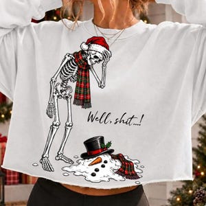 May include: A white long-sleeve sweatshirt featuring a graphic of a skeleton wearing a red Santa hat and a red and green plaid scarf. The skeleton stands over a melted snowman with a black top hat and carrot nose. The text reads, Well, shit....