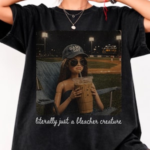 Peut inclure: T-shirt noir avec une poupée portant des lunettes de soleil et une casquette "Game Day", tenant un grand café glacé. La poupée est assise dans une chaise pliante avec un terrain de baseball en arrière-plan. Le texte "literally just a bleacher creature" est en bas.
