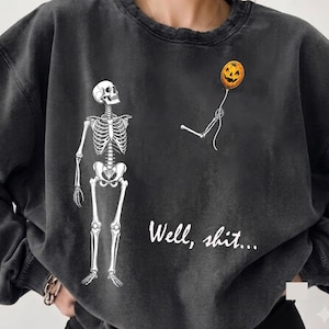 Könnte beinhalten: Ein anthrazitfarbenes Sweatshirt mit Rundhalsausschnitt und einem weißen Skelett-Motiv. Das Skelett steht links, während ein abgetrennter Skelettarm rechts einen orangefarbenen Kürbisballon hält. Der Text "Well, shit..." ist in Weiß gedruckt.