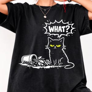 Könnte beinhalten: Schwarzes T-Shirt mit einer weißen Grafik einer grimmigen schwarzen Katze mit gelben Augen und dem Wort "WHAT?" in einer Sprechblase. Neben der Katze steht ein umgestürzter Blumentopf.