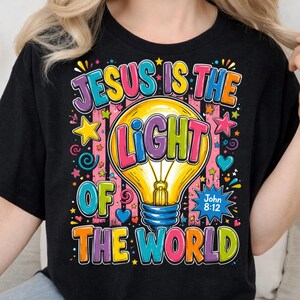 May include: A black t-shirt featuring a colorful graphic of a yellow lightbulb. The text reads "JESUS IS THE LIGHT OF THE WORLD" in multi-colored block letters, with "John 8:12" printed in a blue starburst shape. The design includes stars and hearts.