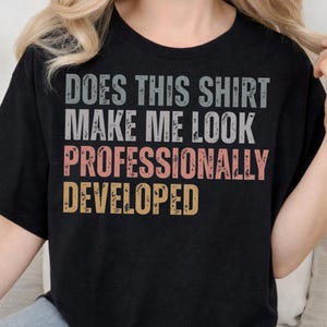 May include: A black short-sleeved t-shirt featuring the distressed-style text DOES THIS SHIRT MAKE ME LOOK PROFESSIONALLY DEVELOPED. The words are printed in stacked lines using grey, light pink, and muted yellow colors with a worn texture.
