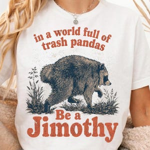 Puede incluir: Una camiseta blanca con un gráfico de un mapache caminando entre la hierba. Sobre el mapache, el texto dice "in a world full of trash pandas" en color óxido. Debajo del mapache, el texto dice "Be a Jimothy" en el mismo color.
