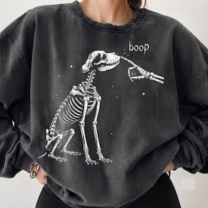 May include: A charcoal grey crewneck sweatshirt featuring a white graphic print of a dog skeleton sitting down while a skeletal finger touches its nose. The word boop is printed in white above the dog's head, surrounded by small white stars.