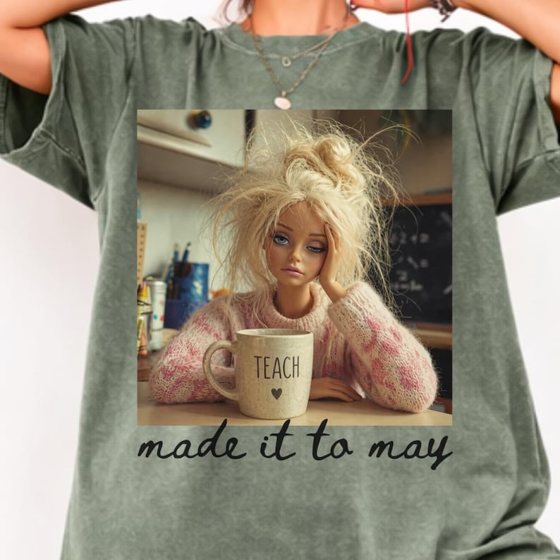 K&ouml;nnte beinhalten: Gr&uuml;nes T-Shirt mit einem Aufdruck, der eine Puppe mit zerzaustem blondem Haar zeigt, die einen rosa Pullover tr&auml;gt und eine Tasse mit der Aufschrift "TEACH" und einem Herz h&auml;lt. Der Text "made it to may" steht darunter.