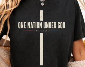 May include: A black t-shirt featuring a large white cross graphic. The text "ONE NATION UNDER GOD" is printed in white across the center, with "SINCE 1776-2026" below it, flanked by a thin red line on the left and a thin blue line on the right.