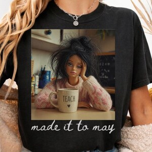 K&ouml;nnte beinhalten: Schwarzes T-Shirt mit einer Grafik einer Puppe mit einer Kaffeetasse mit der Aufschrift "TEACH" und einem Herz. Der Text "made it to may" steht darunter. Die Puppe tr&auml;gt einen rosa-wei&szlig;en Pullover.