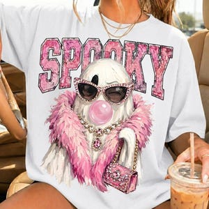 May include: A white t-shirt featuring a graphic of a ghost wearing pink sunglasses, a pink feather boa, a jeweled necklace, and holding a pink handbag. Above the ghost, the word SPOOKY is printed in large, pink, textured block letters.