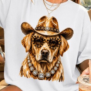 May include: A white t-shirt featuring a graphic print of a golden dog wearing a brown cowboy hat with a studded band, leopard print sunglasses, and a chunky silver necklace with square crystals. The dog has golden fur and a detailed, textured look.