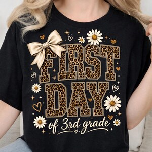 May include: A black t-shirt featuring a large graphic with the text FIRST DAY of 3rd grade. The letters are filled with a brown and black leopard print pattern. A large beige bow, white daisy flowers, and small gold hearts decorate the design.