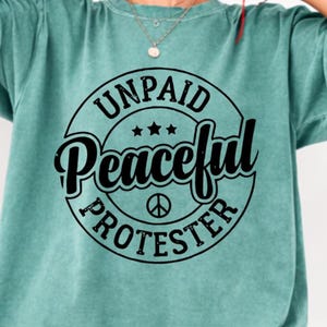 May include: A teal-colored t-shirt with a circular graphic. The graphic reads "UNPAID Peaceful PROTESTER" in a black, bold font, with three stars and a peace symbol. The shirt has short sleeves and a crew neck.