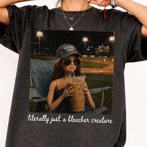 May include: A charcoal gray t-shirt featuring a graphic print of a doll wearing a 'GAME DAY' baseball cap and sunglasses while sitting in a folding chair holding an iced coffee at a baseball field, with the text 'literally just a bleacher creature'.