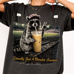 Puede incluir: Camiseta gris oscuro con un mapache con gafas de sol y una gorra "Game Day", sosteniendo una bebida con una pajita y haciendo el signo de la paz. El texto "Literally Just A Bleacher Creature" está debajo.