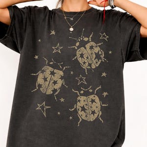 Puede incluir: Una camiseta de manga corta en color negro carbón con un estampado gráfico de tres escarabajos en tono dorado apagado. Cada escarabajo tiene una apariencia texturizada con recortes en forma de estrella en el dorso. Varias estrellas pequeñas dispersas.