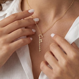 Könnte beinhalten: Eine goldfarbene Halskette mit dem Namen "Catherine" in zarter Schreibschrift. Der Anhänger hängt an einer feinen Kette. Die Halskette wird über einem weißen Hemd getragen.