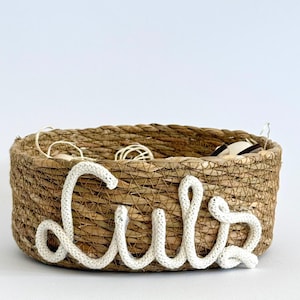 Könnte beinhalten: Ein runder, gewebter Korb aus Naturfasern. Das Wort "Lulu" ist in weißem, strukturiertem Garn auf der Vorderseite geschrieben. Der Korb enthält kleine, weiße Objekte.