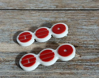 May include: Two white and red contact lens cases. Each case has three connected sections: two oval shapes and one circle. The oval shapes have red horizontal lines, and the circle has a red center. The cases are on a wooden surface.