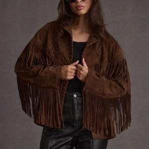 May include: A brown suede jacket with long fringe detailing. The jacket has a classic collar and is worn over a black top and black leather trousers. The model is wearing black sunglasses.
