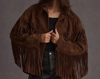 May include: A brown suede jacket with long fringe detailing. The jacket has a classic collar and is worn over a black top and black leather trousers. The model is wearing black sunglasses.