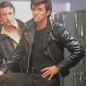 Puede incluir: Una chaqueta de motorista de cuero negro con cremalleras y broches plateados, usada sobre una camiseta negra lisa. La chaqueta presenta un cuello ancho con muescas, charreteras en los hombros y una cintura con cinturón, combinada con pantalones negros.