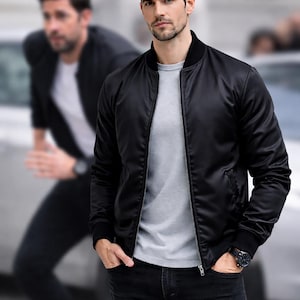 May include: A black bomber jacket with a black ribbed collar and cuffs. The jacket is zipped up and worn over a grey t-shirt and black jeans. A watch is visible on the wrist. Another person is blurred in the background.
