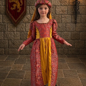 Puede incluir: Una joven con un vestido medieval rojo y dorado y una diadema a juego. El vestido presenta un patrón detallado y mangas largas. El fondo incluye una pared de piedra, una pancarta con un emblema de león y una antorcha.