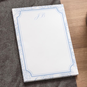 May include: A rectangular notepad with a white background and a light blue floral border. The border frames the notepad with a decorative design. The initials "JB" are written in a light blue script font in the upper center of the notepad.