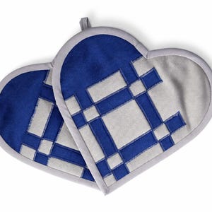 May include: Two heart-shaped pot holders. Each pot holder has a blue top section and a white and blue geometric design. The edges are gray, and each has a small gray loop for hanging.