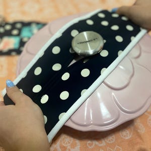 May include: A pink, scalloped-edge serving dish with a navy blue and white polka dot handle cover. A small, silver metal lid is centered on the dish. A matching pot holder is visible in the background.