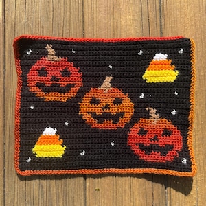Puede incluir: Una pieza rectangular de ganchillo con fondo negro, tres calabazas de Halloween naranjas y dos dulces de maíz. El diseño incluye pequeños detalles bordados en blanco y está rematado con un borde naranja vibrante en los extremos.