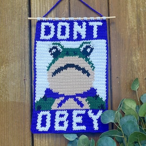 Puede incluir: Un tapiz de pared hecho a mano con ganchillo con las palabras "DONT OBEY" en letras blancas sobre un fondo azul. La obra de arte presenta una rana con una expresión triste, una pajarita y detalles en verde y azul. El tapiz está suspendido de una varilla de madera.