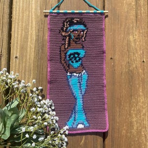 May include: A crocheted wall hanging depicting a woman in blue clothing. The artwork is framed with a pink border and suspended from a wooden dowel with a teal cord. The background is a wooden surface. The piece is handmade.