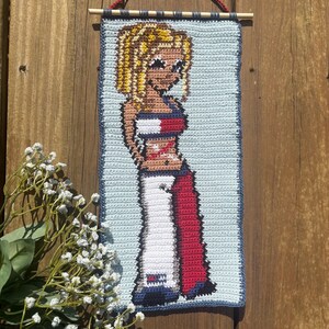 May include: Crocheted wall hanging depicting a blonde woman in a red, white, and blue outfit. The artwork has a light blue border and a wooden dowel with a red string for hanging. The background is a wooden surface.