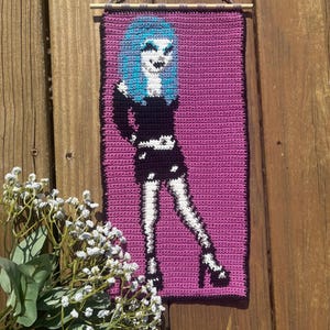 May include: A handmade crochet wall hanging depicting a figure with blue hair, a black dress, and white legs and shoes, against a purple backdrop. The artwork is hung from a wooden dowel with a grey cord.