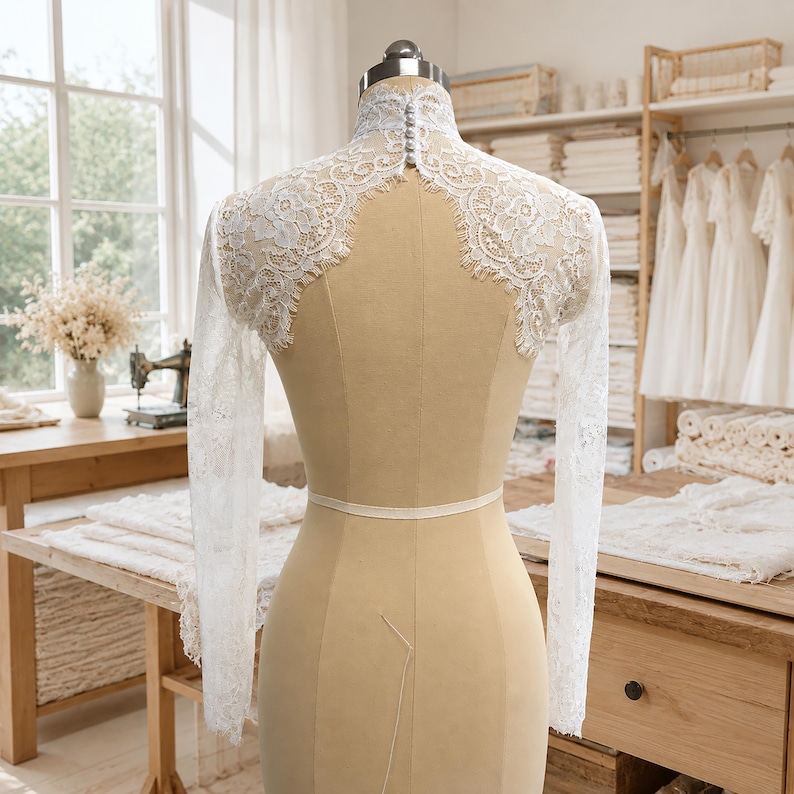 May include: A white lace bolero with long sleeves and a high neck, featuring intricate floral patterns and a vertical row of small round buttons at the back of the collar, displayed on a beige dress form in a bright studio setting.