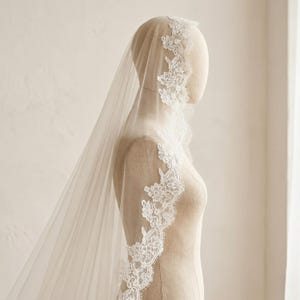 May include: A long, sheer ivory wedding veil draped over a mannequin. The veil features an intricate white floral lace trim along the edges, extending from the top of the head down to the lower back, creating a soft, layered appearance.