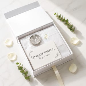 Puede incluir: Una caja de regalo blanca con tapa magn&eacute;tica que contiene papel de seda blanco, un peque&ntilde;o objeto circular envuelto en pl&aacute;stico de burbujas y una tarjeta rectangular con el texto ENDLESS THANKS For your order impreso en negro.