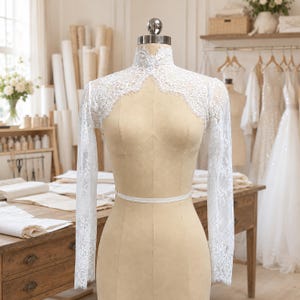 May include: A white lace bolero with long sleeves and a high mock neck collar, featuring intricate floral patterns and scalloped edges. The garment is displayed on a beige dress form, showcasing the delicate lace texture and fitted design.