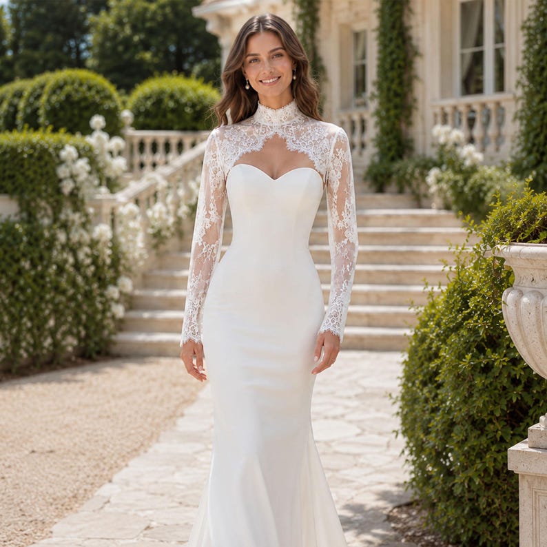 May include: A floor-length white gown featuring a sweetheart neckline and long, sheer lace sleeves that extend into a high-neck lace bodice overlay. The dress has a fitted silhouette that flares slightly toward the hemline.