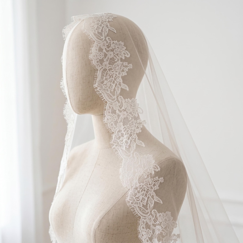 Puede incluir: Un velo de novia blanco y transparente con un intrincado borde de encaje floral. El delicado patr&oacute;n de encaje incluye flores y hojas detalladas con un acabado festoneado, colocado sobre la cabeza y los hombros de un maniqu&iacute;.