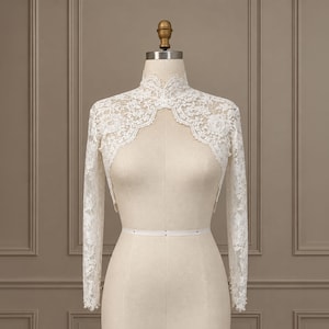 May include: A white lace bolero jacket featuring a high neckline, long sleeves, and intricate floral patterns. The garment is displayed on a mannequin, showcasing a scalloped edge along the front opening and small pearl buttons at the cuffs.