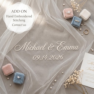 Puede incluir: Un arreglo de boda con texto en cursiva blanca que dice Michael & Emma 09.14.2026, rodeado de tela blanca transparente, cajas de terciopelo para anillos en rosa y azul, peque&ntilde;as perlas blancas, flores secas y una tarjeta de invitaci&oacute;n.